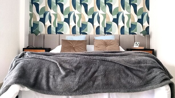 Un lit bien fait, avec une couverture grise, deux oreillers et une tête de lit ornée d’un motif géométrique.
