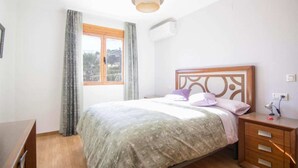 Quarto com cama, mesa de cabeceira, cômoda de madeira, janela com cortinas e ar condicionado.