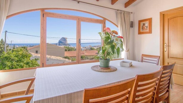 Uma área de refeições com uma mesa posta para quatro pessoas, uma planta em vaso e vista para o mar e prédios.