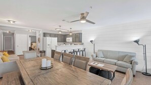 A modern kitchen with dark cabinets, a white refrigerator, and a microwave. There is a dining area with a wooden table and chairs, and a gray sofa with a floor lamp.