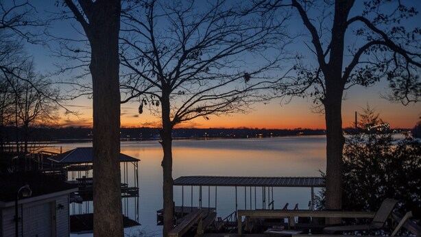 Ein Steg am Seeufer mit einer Holzterrasse und Liegestühlen, mit Blick auf ruhiges Wasser bei Sonnenuntergang.