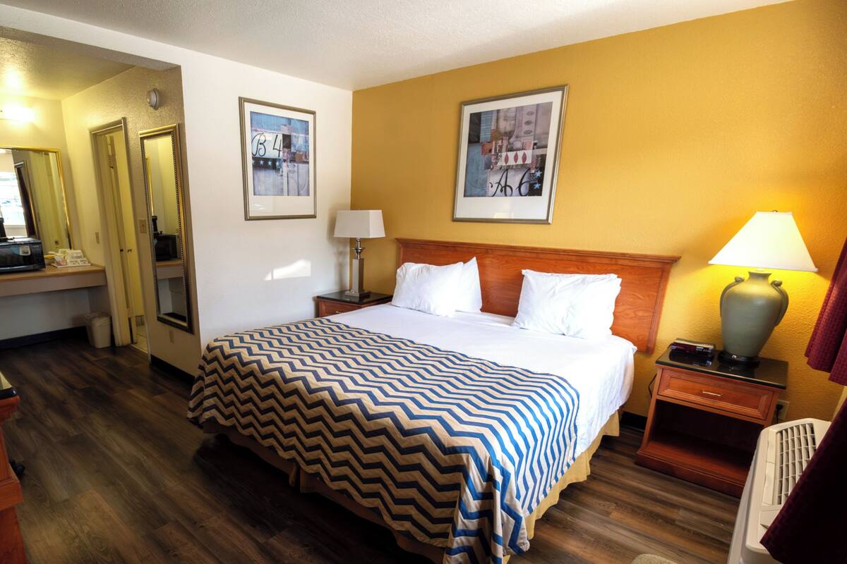 A hotel room with a large bed, wooden floor, a desk with a chair, a lamp, and two framed pictures on the wall.