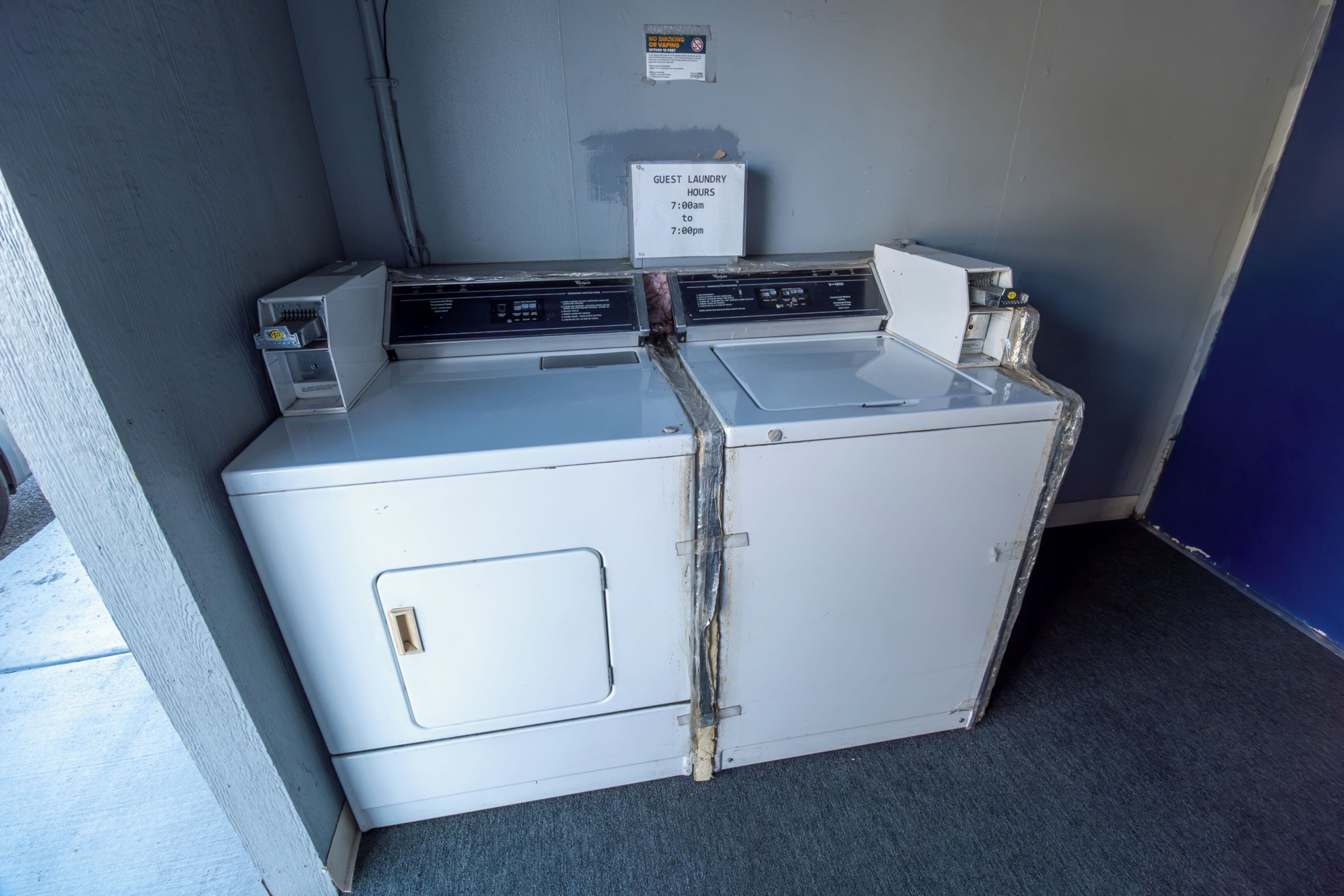 Een compacte, witte wasmachine, bevestigd aan de muur, met een bord waarop de openingstijden van de wasruimte staan.