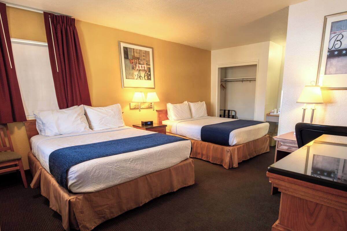 A hotel room with two beds, a desk, a chair, a wardrobe, and a painting on the wall.