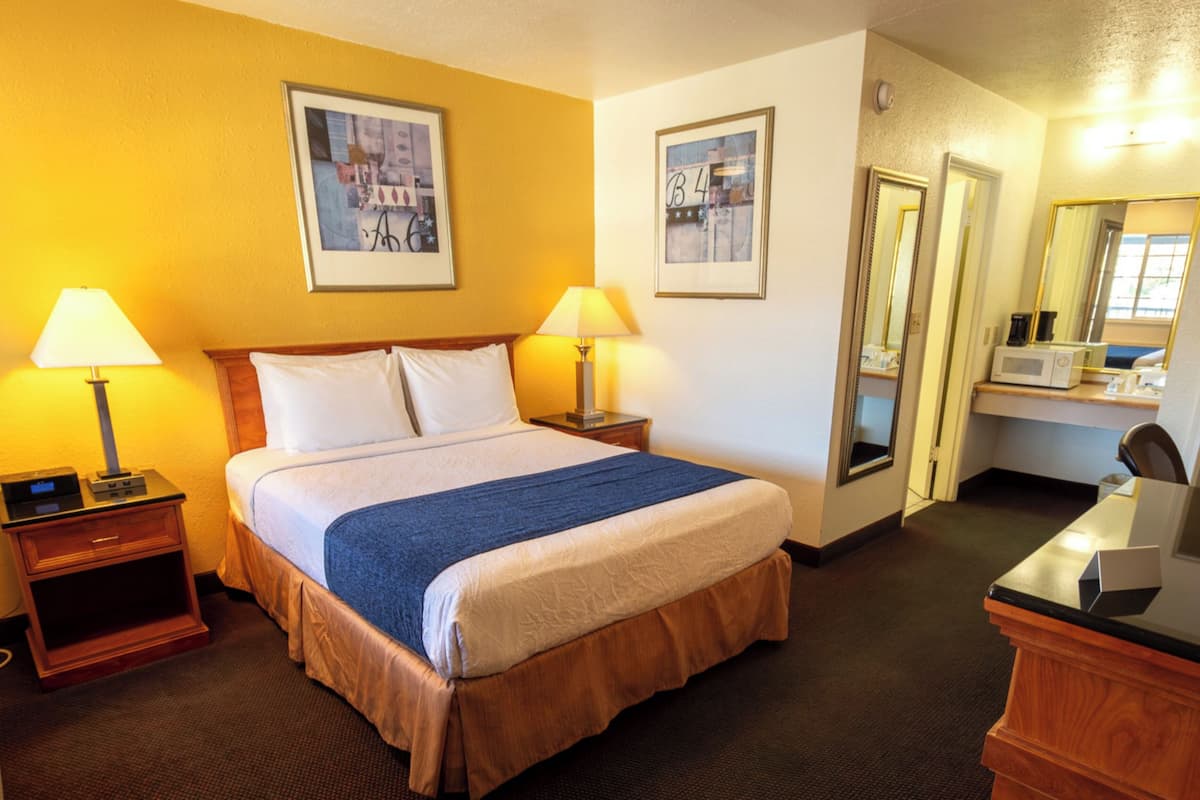 A hotel room with a bed, two bedside lamps, a nightstand, a desk with a chair, a mirror, and framed pictures on the walls.