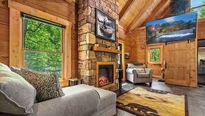 A cozy log cabin interior with a stone fireplace, a large window overlooking greenery, a comfortable sofa, and a television mounted on the wall. - Timber View Hideaway – Cozy Mountain Retreat Between Gatlinburg & Pigeon Forge! (Sevierville)