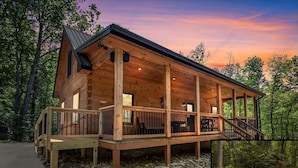 A wooden cabin with a covered porch, stone foundation, and a fire pit area.