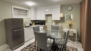 A compact kitchen with white cabinets, a refrigerator, a microwave, and a coffee maker. There is a dining table with four chairs and a patterned rug on the floor.