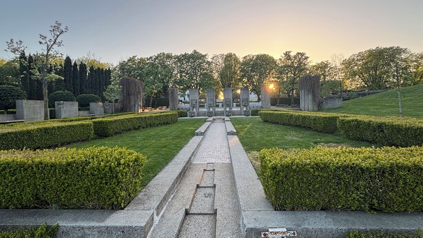 A formal garden with a central pathway leading to a monument, surrounded by neatly trimmed hedges and trees.