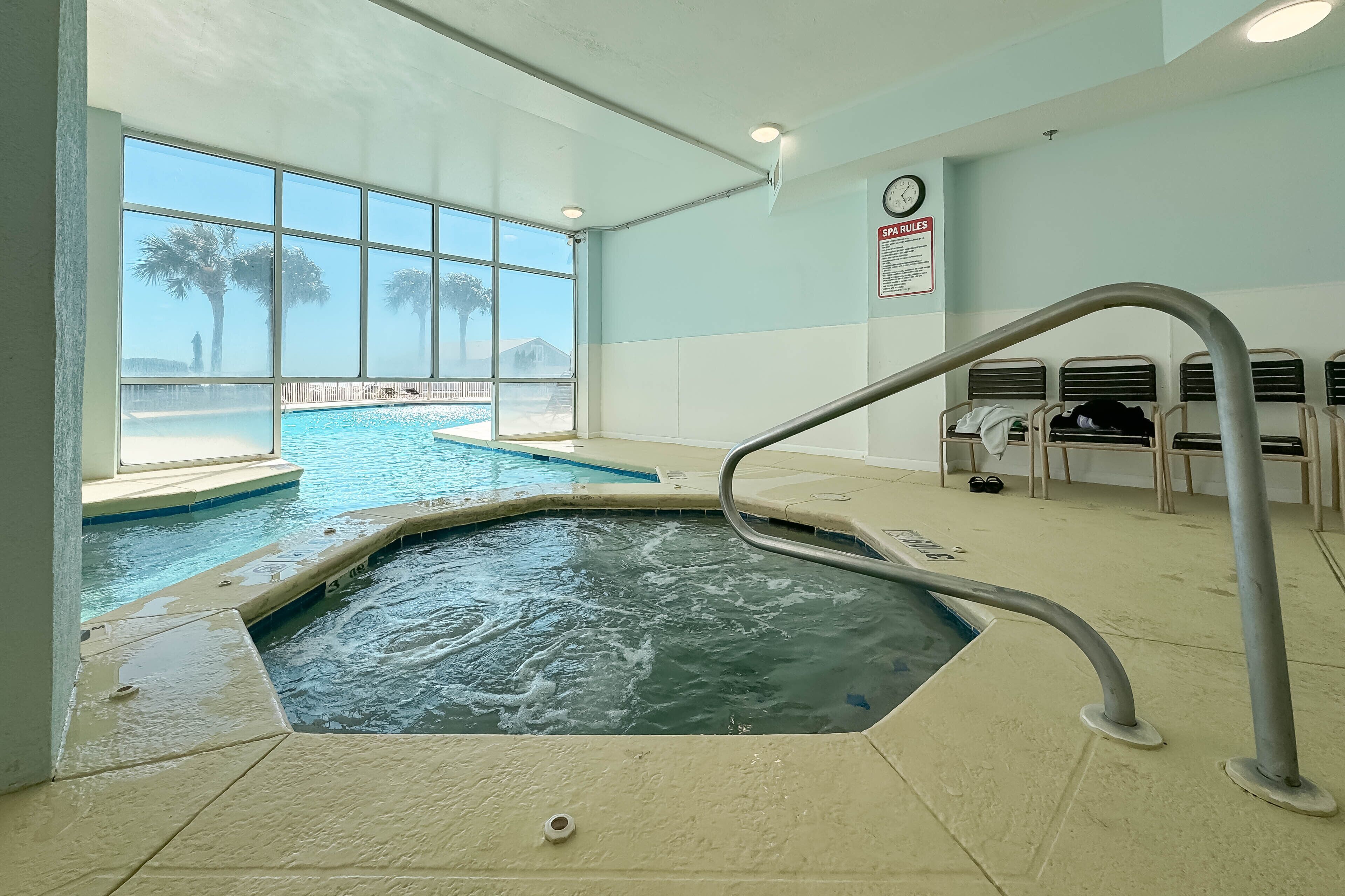 An indoor swimming pool with a view of palm trees and a beach, a spa rules sign, and a clock on the wall.