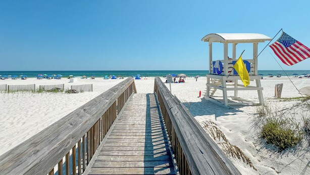 A wooden boardwalk leading to a lifeguard station with an American flag and a yellow flag. - PRIVATE HOME WITH BEACH ACCESS AND POOLS! (Gulf Shores)