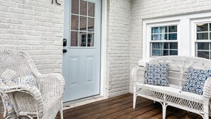 Fachada de ladrillo blanco con una puerta azul claro, dos sillas de mimbre y un sofá de mimbre con cojines estampados.