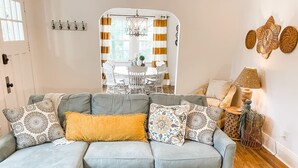 Uma sala de estar acolhedora com um sofá seccional cinza, almofadas estampadas, um cobertor amarelo e uma mesa de centro rústica. Uma área de jantar com uma mesa e cadeiras brancas é visível por uma janela com cortinas listradas.