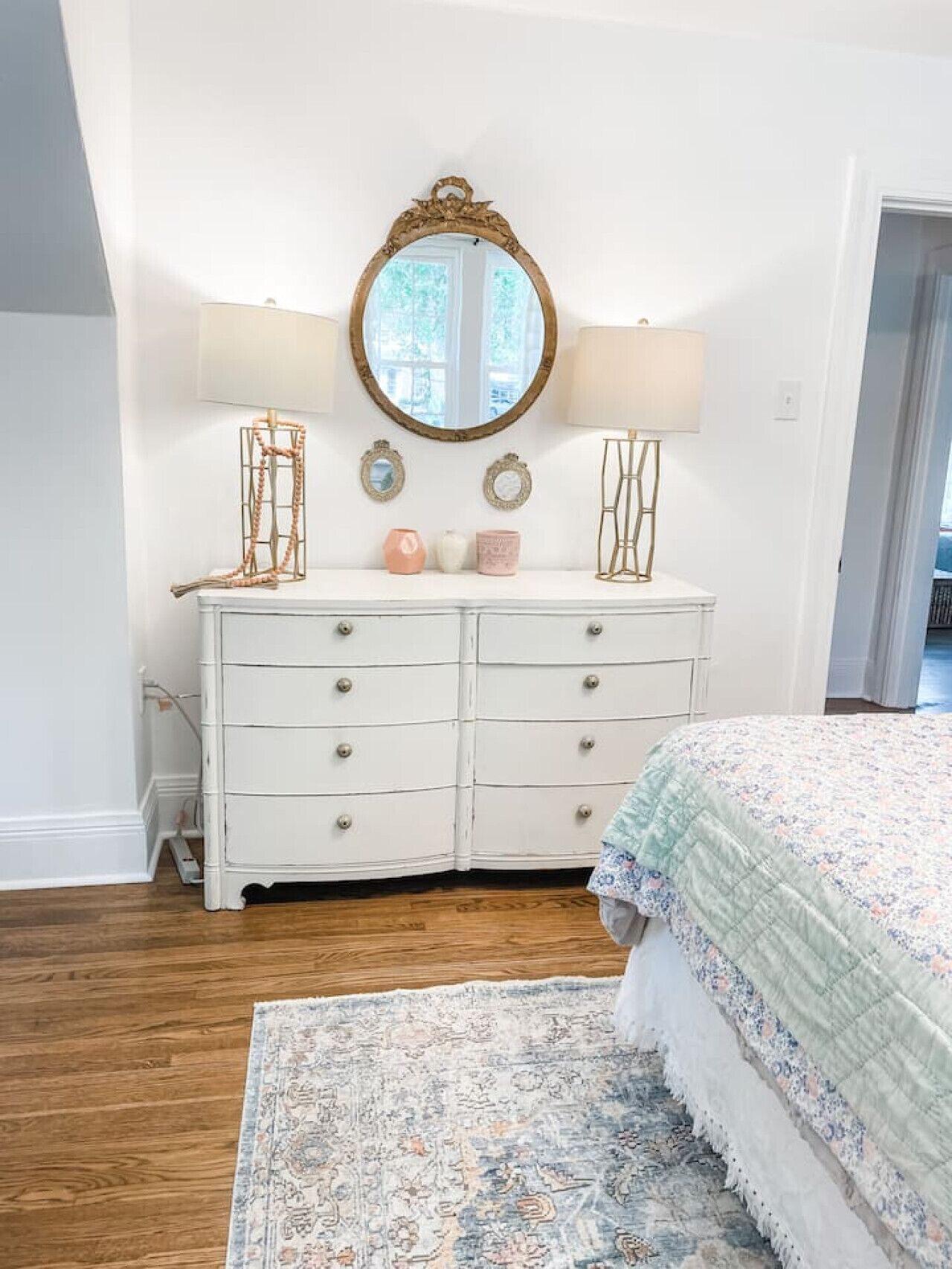 A bedroom with a white dresser, a round mirror, two wall sconces, and a patterned rug.