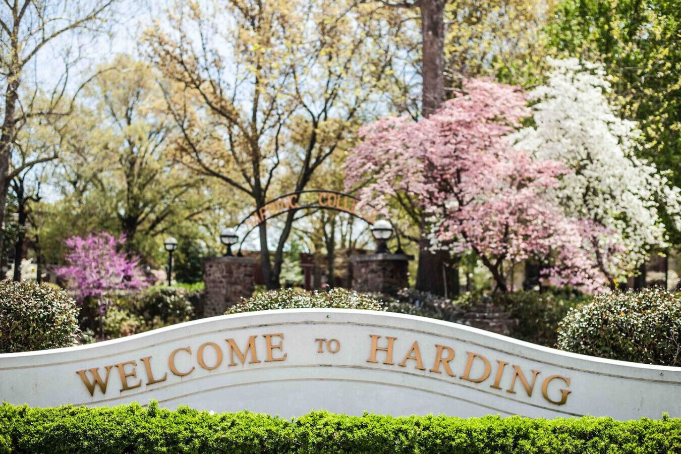 A welcome sign with "WELCOME TO HARDING" in gold lettering on a white background, surrounded by lush greenery and flowering trees.
