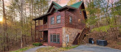 A rustic cabin with a stone foundation, a wooden deck, and a staircase leading to the main entrance.