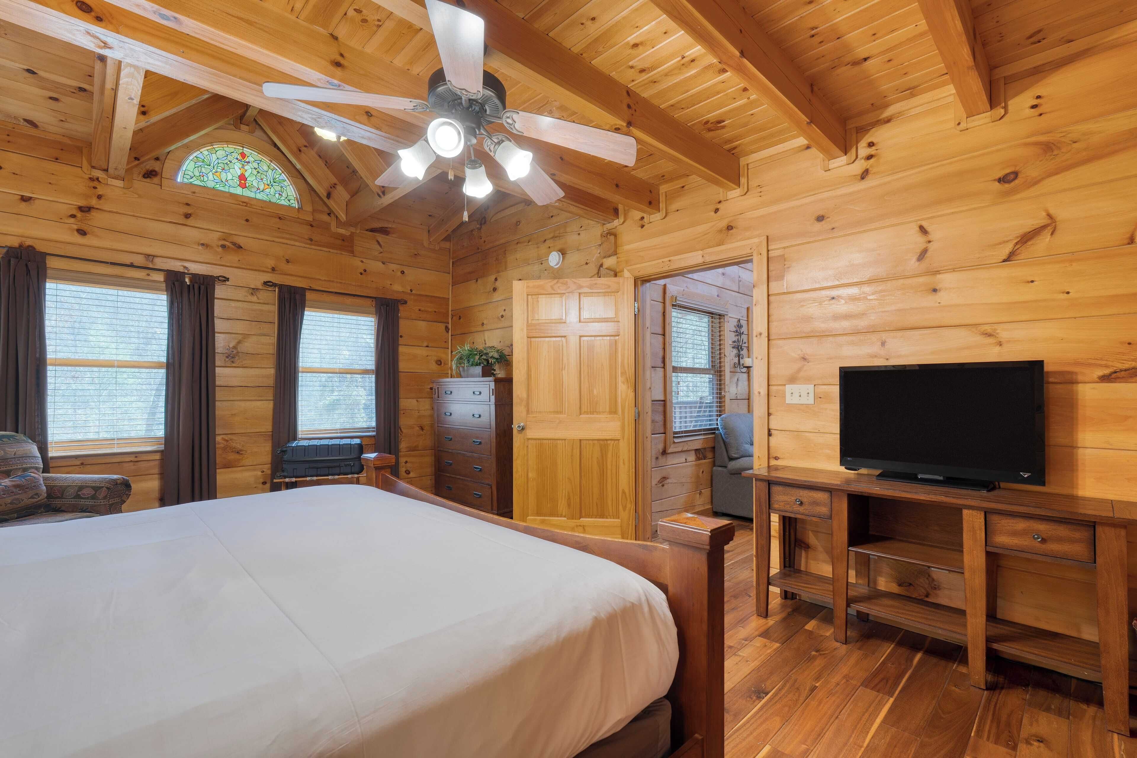 Una habitación de cabaña de madera con una cama, un ventilador de techo, un televisor y una ventana con cortinas.
