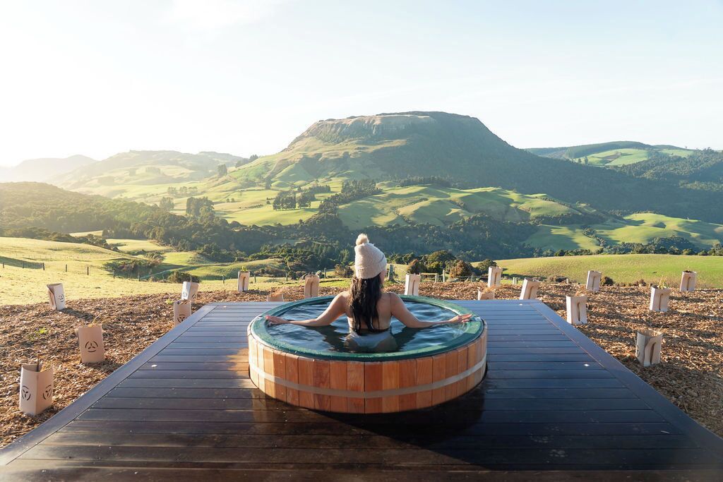 A person sitting in a wooden hot tub on a wooden deck with a scenic view of rolling hills and a mountain in the background.