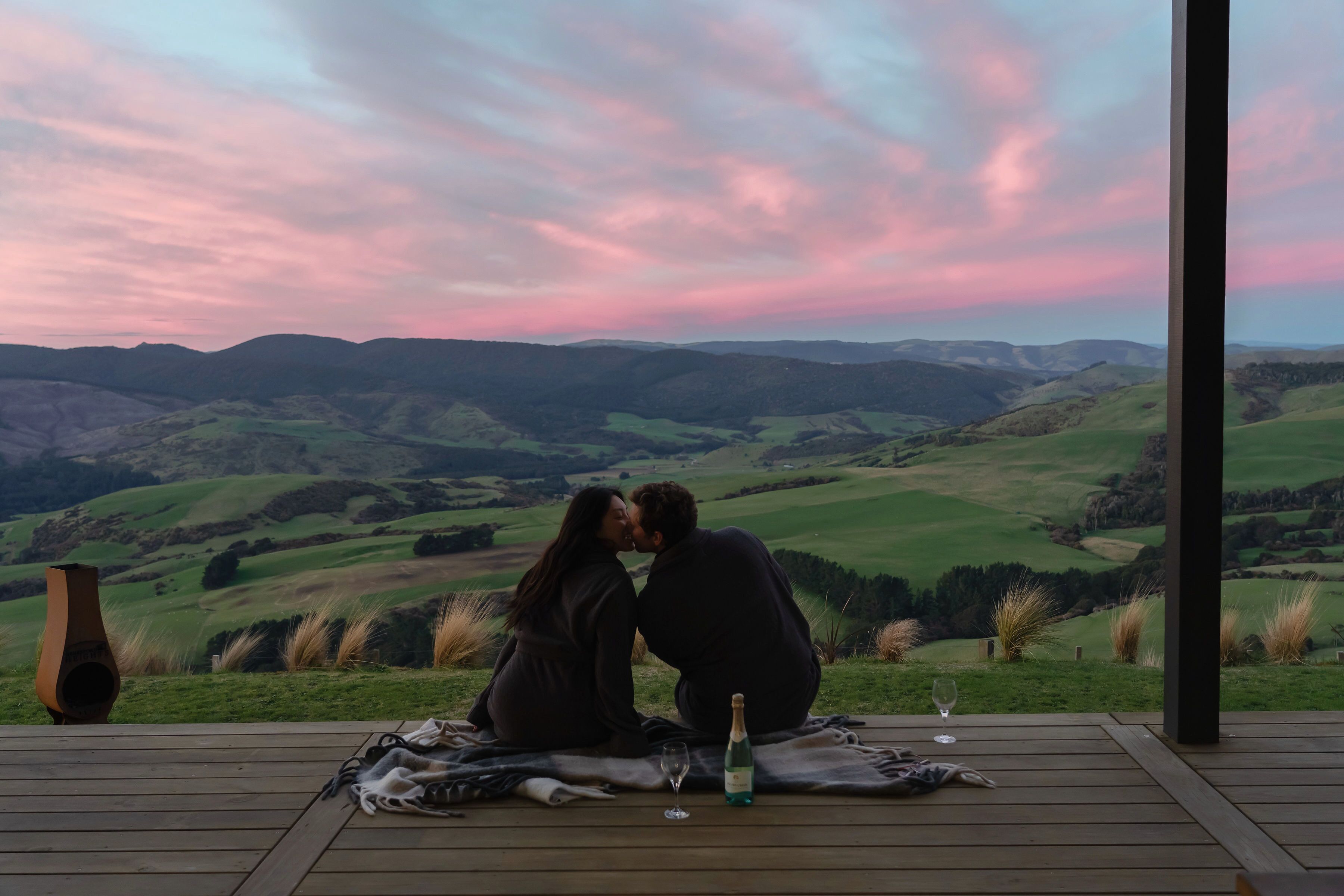 A couple on a wooden deck overlooking a valley with a sunset sky.