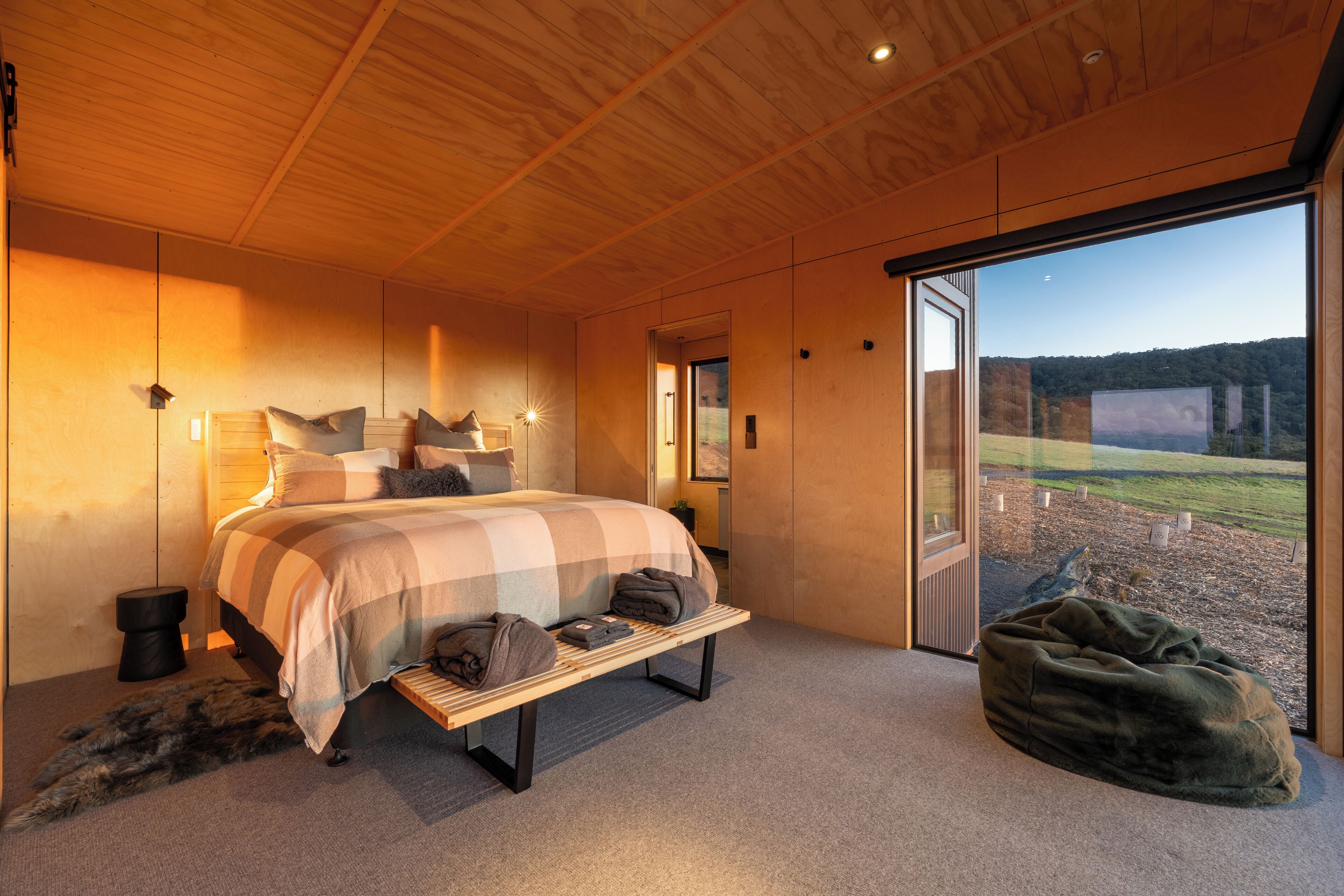 Un dormitorio con una cama grande, un banco, vistas a través de un ventanal y un ambiente acogedor.