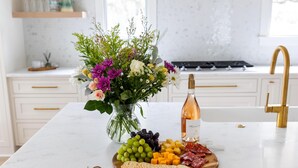 A kitchen with a marble countertop, a wooden cutting board with cheese, grapes, and crackers, a bottle of wine, and a vase of flowers. - Newly Built IOP Home – Pool, Ping Pong, Walk to Beach, Shops & Restaurants (Isle Of Palms)