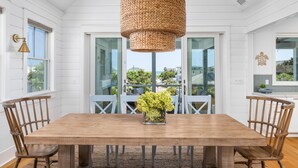 Uma área de jantar com mesa de madeira, cadeiras, luminária trançada pendente e vista para o exterior através de portas de vidro.