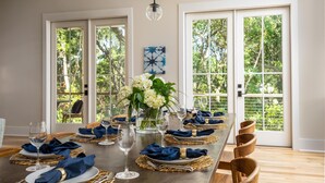 Une salle à manger avec une longue table dressée pour six personnes, de grandes fenêtres et un lustre.