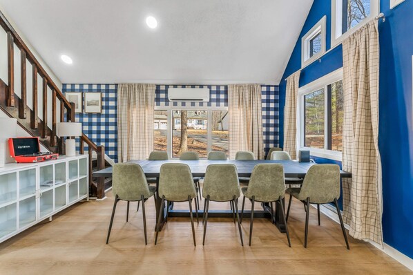 A dining area with a long table and six chairs, a staircase with wooden railings, and a view of a house through a window.