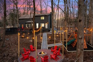 A cabin with a fire pit and red chairs in a wooded area, surrounded by trees and a playground.