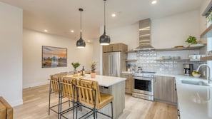Une cuisine moderne dotée d’un îlot central, d’appareils électroménagers en acier inoxydable et de luminaires suspendus.