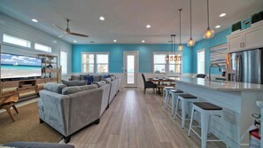 A spacious living area with a large sofa, a television, and a kitchen area with a refrigerator and bar stools. - Sleeps 22! 2 Homes Priv Pool, Putting Green, Bar, Deck, Prime Port A Location (Port Aransas)