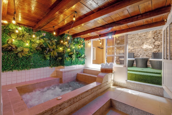 Un área de spa con hidromasaje, techo de madera, pared verde con plantas, muebles de mimbre y pared de piedra.
