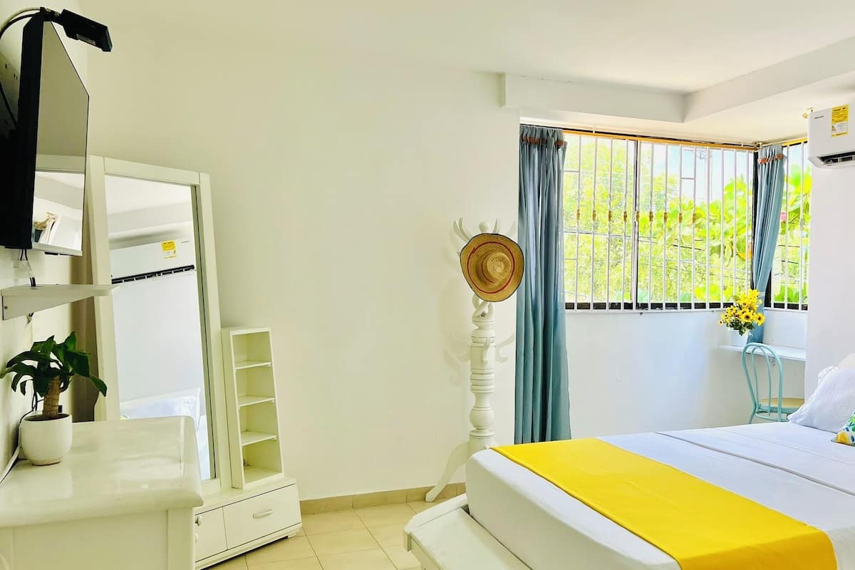 A bedroom with a bed, a bedside table, a mirror, a window with curtains, a potted plant, and an air conditioning unit.