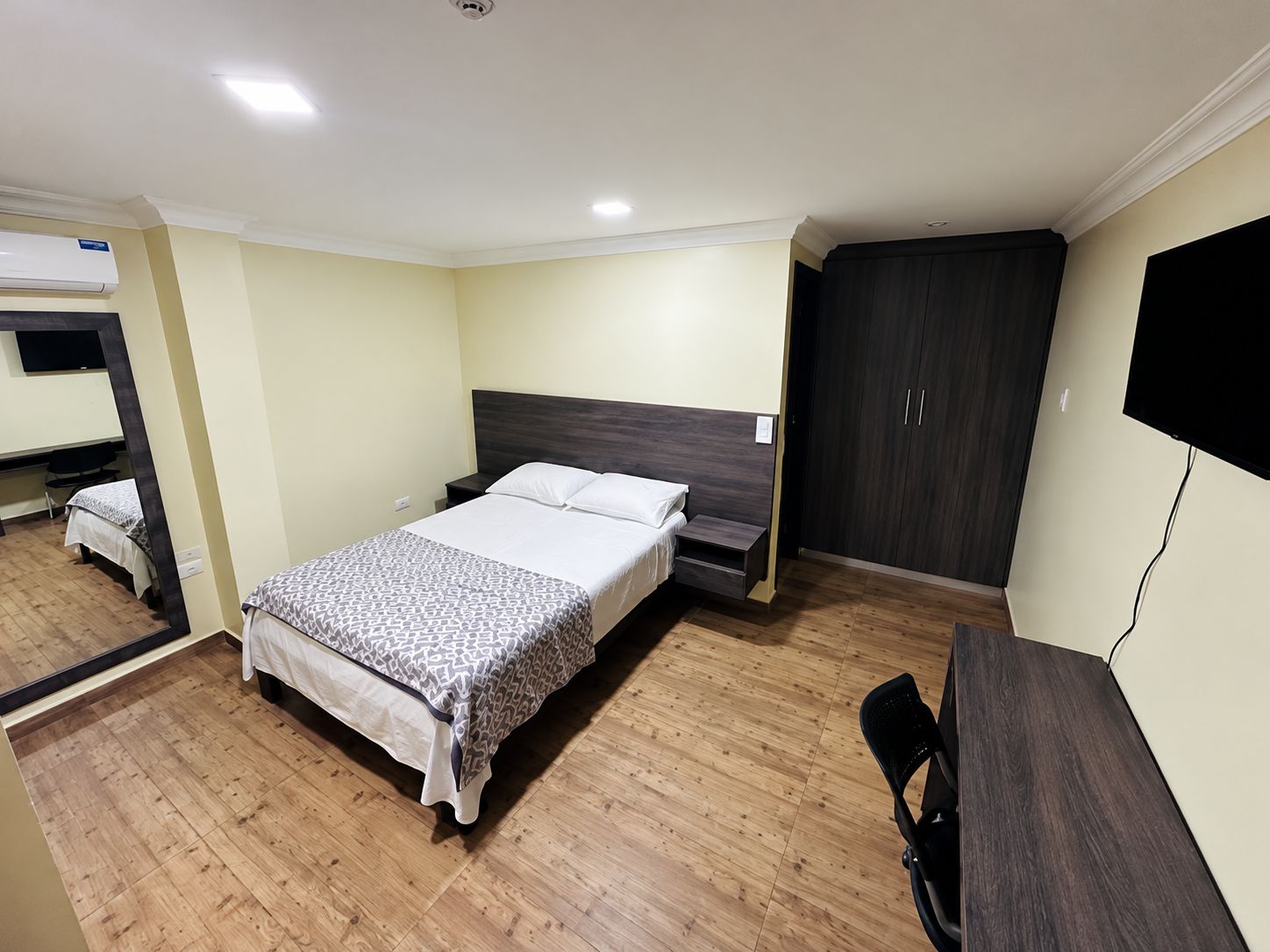 Quarto de hotel com cama, escrivaninha, guarda-roupa e televisão.