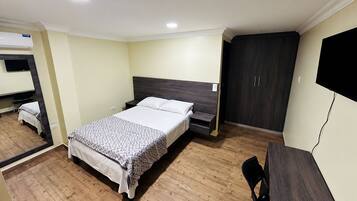 Quarto de hotel com cama, escrivaninha, guarda-roupa e televisão.