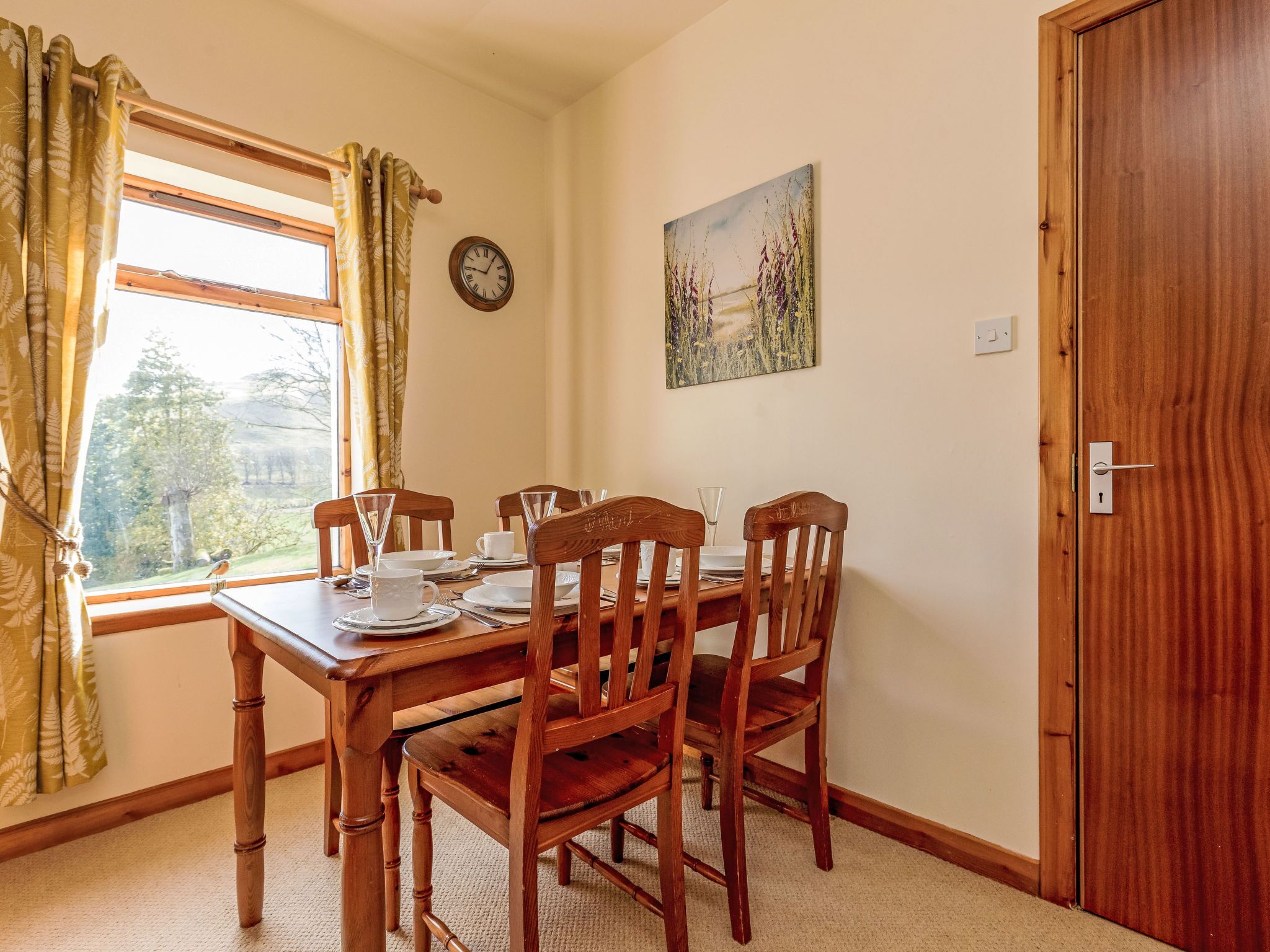 A dining area with a wooden table and chairs, a window with curtains, a clock, and a painting on the wall.