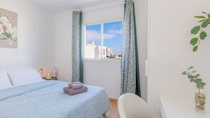A bedroom with a bed, a bedside table with a lamp, a window with curtains, and a view of buildings outside.