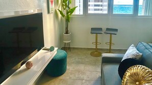 Una sala de estar con televisor, planta, dos taburetes altos, sofá y vista al océano a través de amplias ventanas.