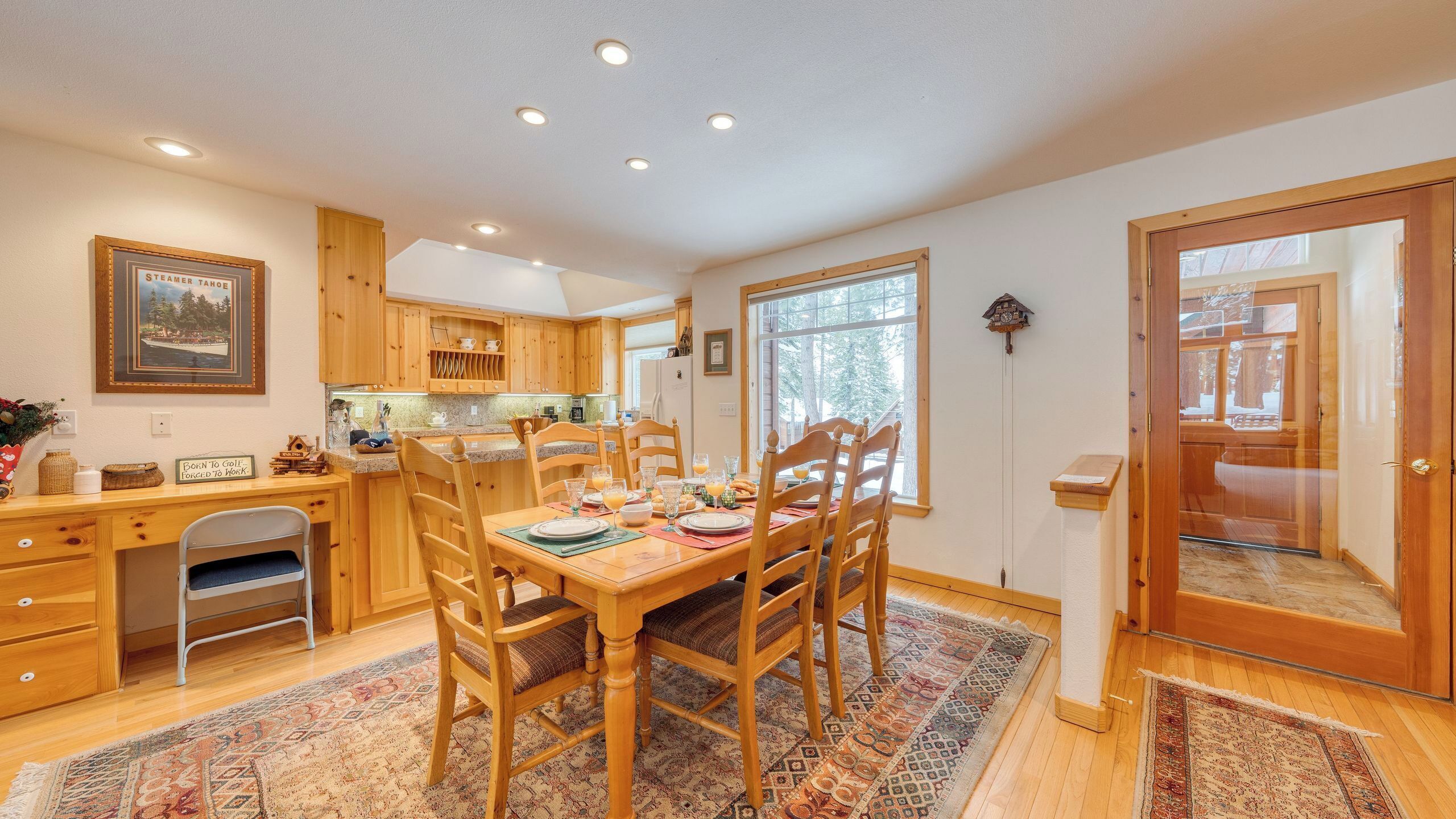 A dining area with wooden furniture, a kitchen in the background, and a large window.