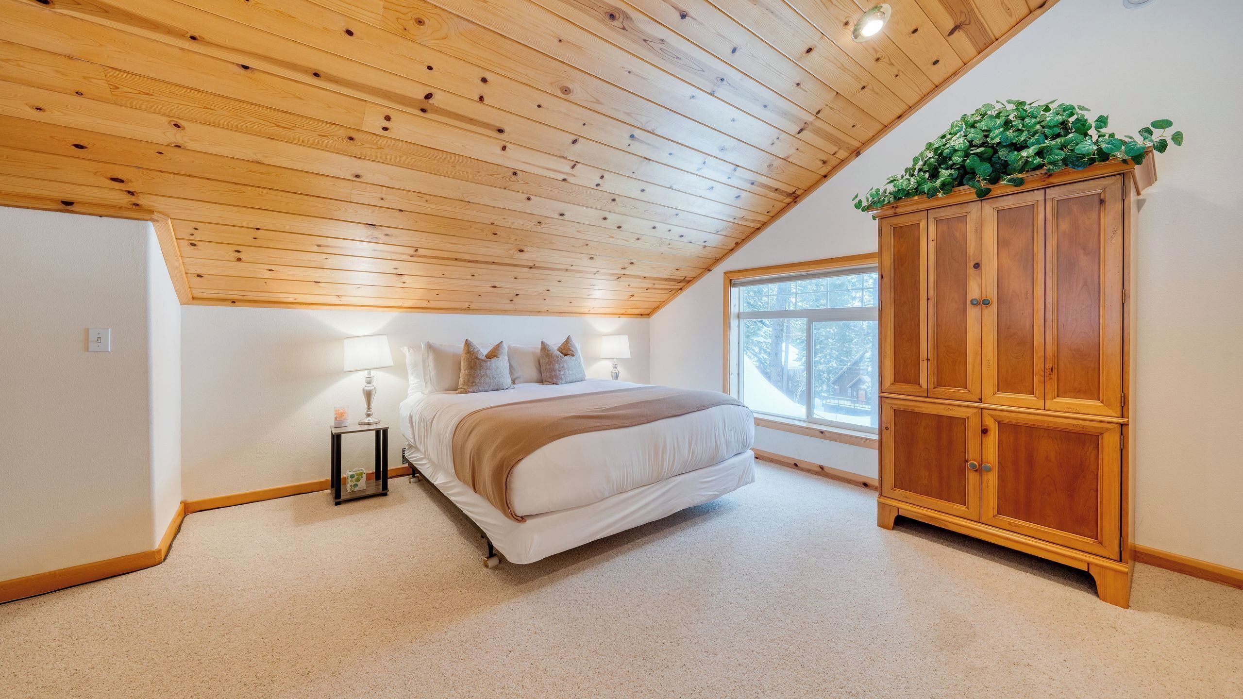 Ein Schlafzimmer mit Holzbalkendecke, einem Bett mit beiger Decke, einem Nachttisch mit Lampe und einem Holzschrank.