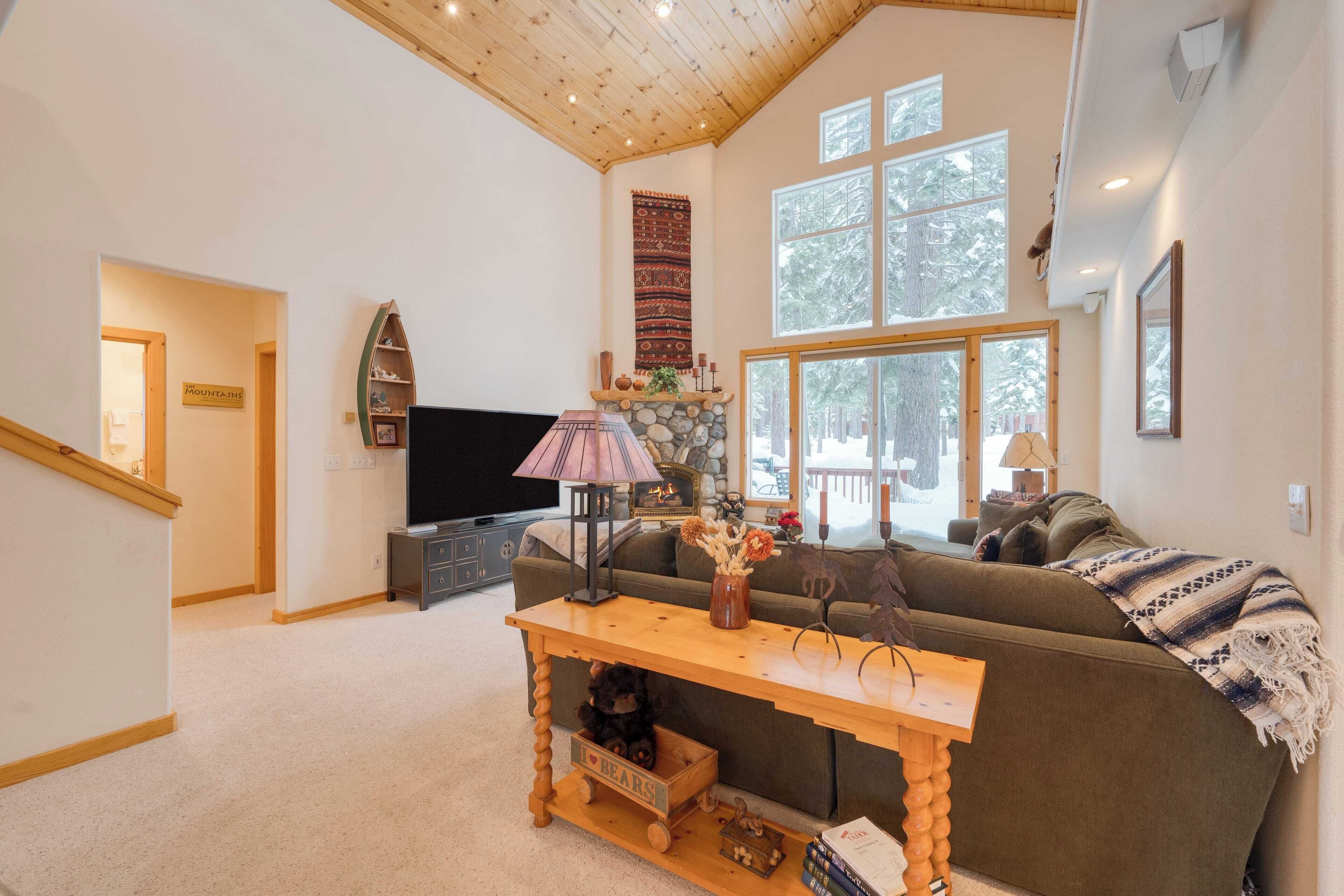 Ein gemütliches Wohnzimmer mit Holzbalkendecke, einem großen Fenster, einer Couch, einem Fernseher und einem Tisch mit Teddybär und Büchern.