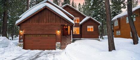 A wooden cabin with a garage, surrounded by snow-covered ground and tall pine trees.