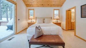 Un dormitorio con una cama grande, techo de madera, dos lámparas de noche, un banco y vista a una terraza nevada.