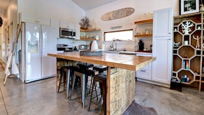 A modern kitchen with white cabinetry, a central island with stools, a microwave, a refrigerator, and a wooden shelf unit with decorative items. - Midnight Eddy @ Water's Edge: Lake front~Hot Tub~Fire pit~Kayaks~more (Fayetteville)
