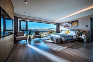 Un dormitorio moderno con una cama grande, un área de descanso con vistas y un paisaje urbano visible a través de amplios ventanales.