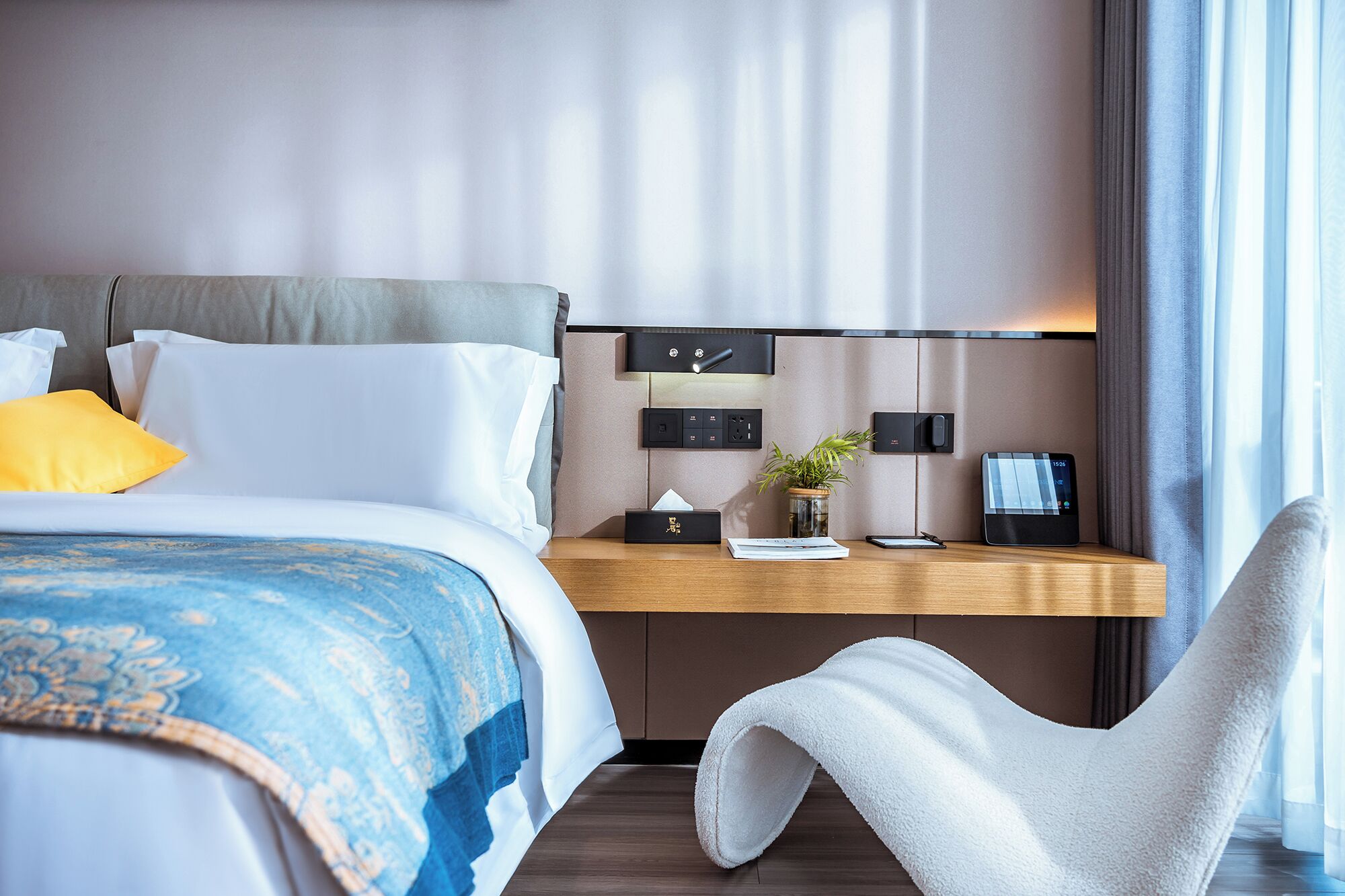 Een moderne hotelkamer met een net opgemaakt bed, een bureau met een laptop, een plant en een stoel.