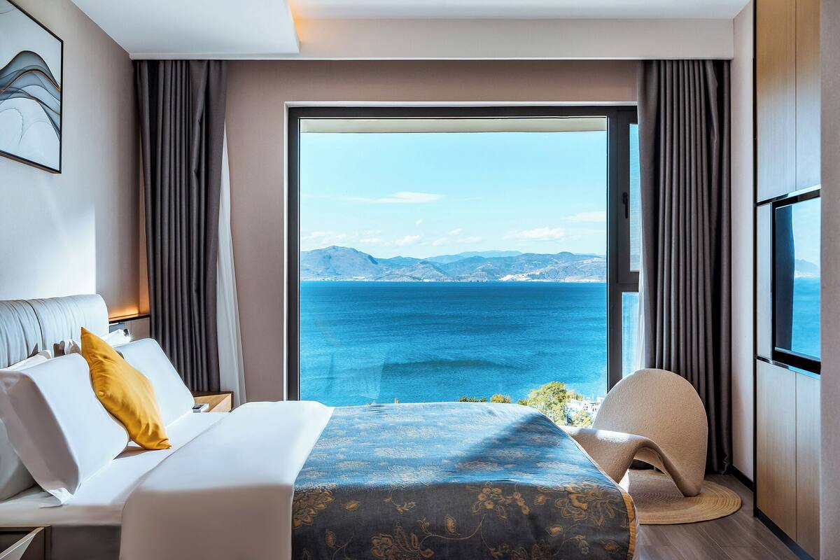 Een hotelkamer met een bed, een stoel, een groot raam met uitzicht op zee en bergen, en een schilderij aan de muur.
