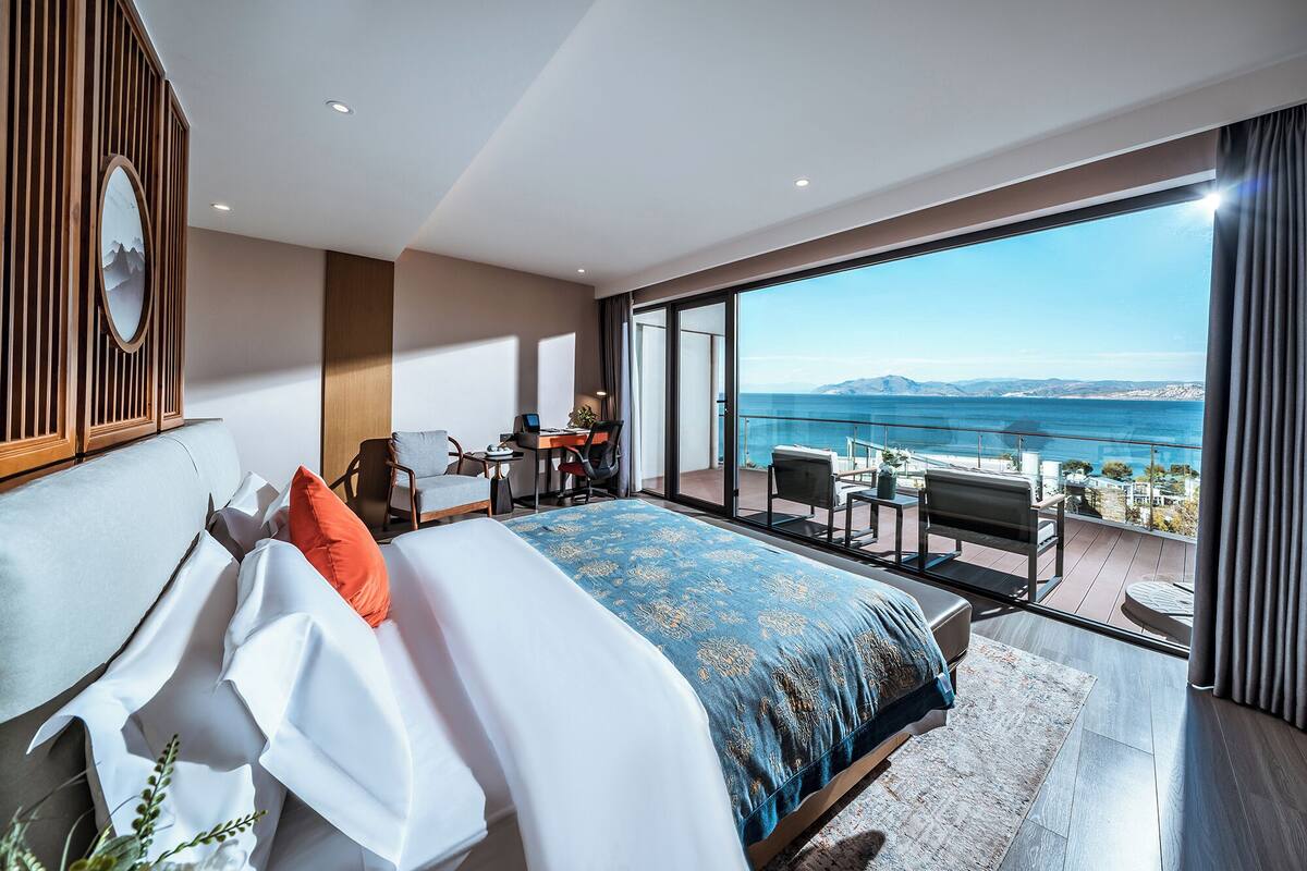 Een moderne hotelkamer met een groot bed, een bureau met een laptop, en een balkon met uitzicht op zee.