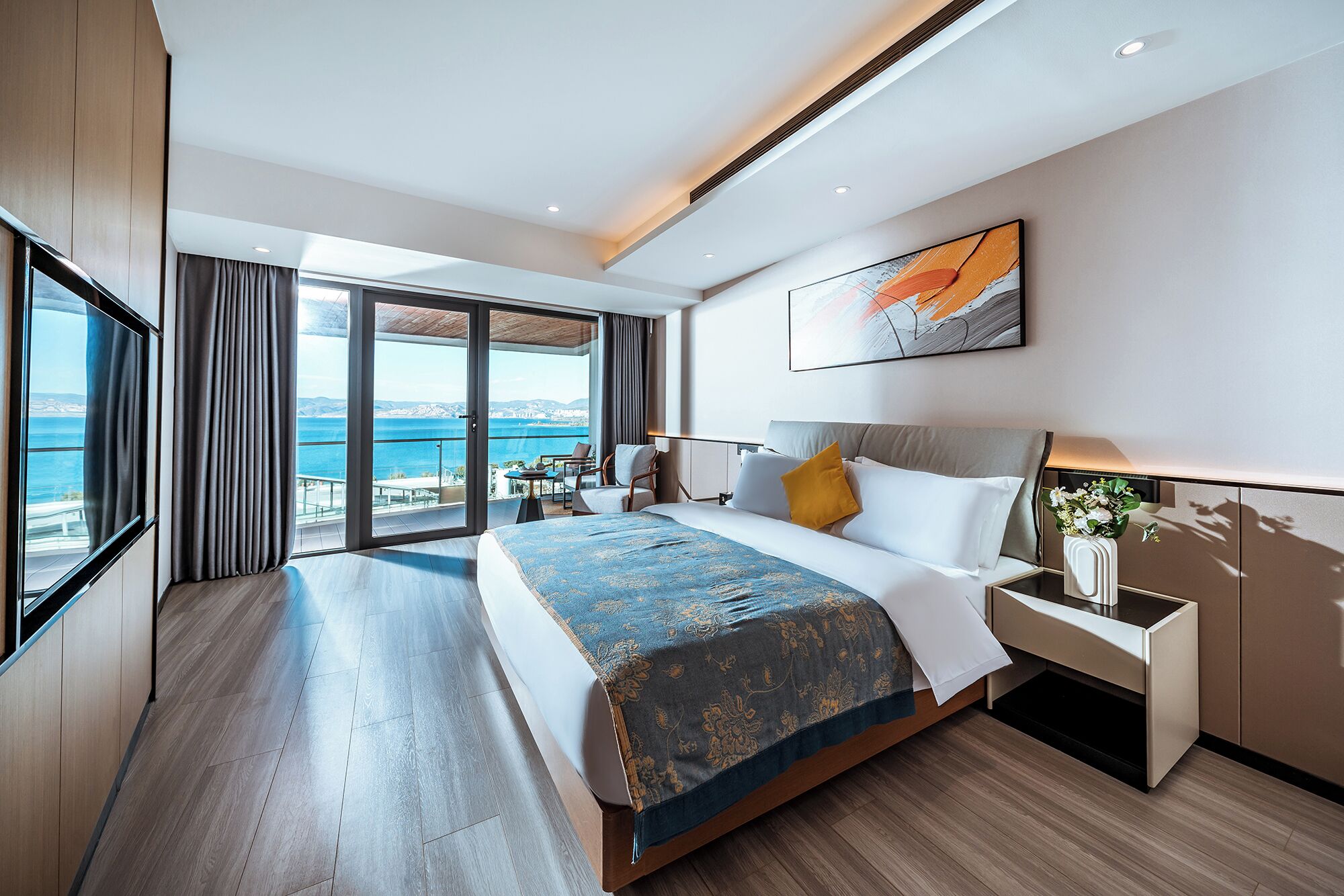 A modern hotel room with a large bed, a bedside table, a nightstand, a view of the sea, and a painting on the wall.