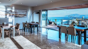 Un espace repas moderne avec une grande table et des chaises, une porte vitrée donnant sur un balcon avec vue sur la mer et les montagnes.